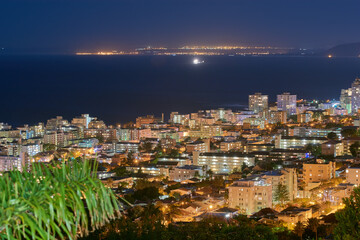 Fototapeta premium Urban city lights against a midnight sky with copy space. Skyline with colorful lighting and the open ocean on the horizon. Modern architectural buildings, vacation hotels in an urban city