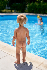 Small naked baby stands by the pool with turquoise water. Back view