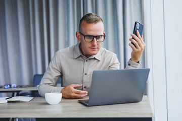 Successful man talking on smartphone and browsing netbook while surfing internet in office at table