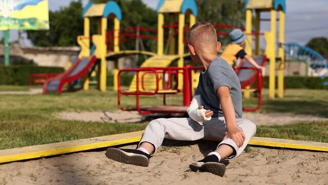 Child with broken hand wrapped in white plaster bandage. Concept of healthcare. The boy holds a broken arm and looking how another kids playing on playground the background. spoiled summer vacation.