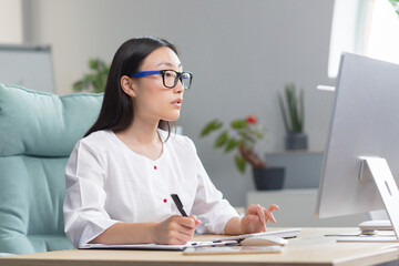 The doctor is online. A young beautiful Asian woman doctor conducts an online consultation. He is sitting in a modern office at a work table, talking on a video call with a patient at a computer.