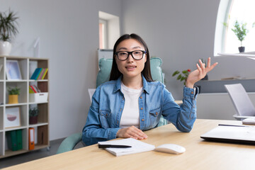 Distance Learning. A young Asian woman is a teacher, conducts online classes, lessons. He looks into the camera, explains, tells. Sitting in glasses and jeans at a table at a computer in a classroom