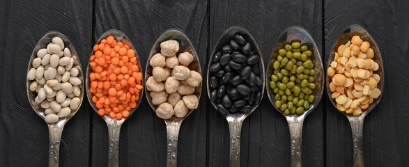 Variety of legumes in old silver spoons on a black wooden background.