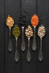 Fototapeta na wymiar Variety of legumes in old silver spoons on a black wooden background.