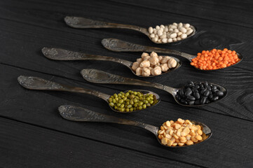 Variety of legumes in old silver spoons on a black wooden background.