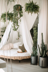 A bright, tropical-style room with a white four-poster bed, white and pink pillows, flowers hanging from the ceiling, and cacti in flowerpots.