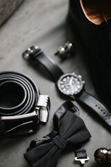 A men's wrist watch, a black butterfly, and a rolled-up leather belt lie on a dark table.