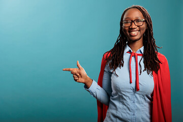 Joyful and confident superhero woman wearing red cape while pointing fingers to left on blue background. Selfless and strong young adult justice defender person posing at camera. Studio shot.