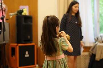 A little girl plays the trumpet back view in class at school with a brunette teacher with long hair...