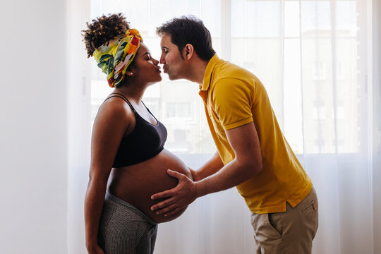 Multiracial pregnant couple kissing in natural light room
