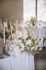 Wedding decor of a banquet table of snow-white color. White roses, peonies, gypsophila, white candles in glass candlesticks.