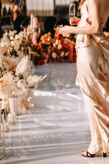 A girl in a beige silk dress that develops in the wind stands and holds a glass of champagne. In the background are a white glossy floor and a decor of flowers and candles.
