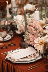 Wedding banquet table decor with pink and white flowers and burning candles on an orange tablecloth. The restaurant menu is on a plate with a silk napkin.