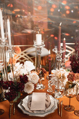 Wedding banquet table decor with pink and white flowers and burning candles on an orange tablecloth. The restaurant menu is on a plate with a silk napkin.
