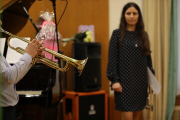 A student playing a golden trumpet in a classroom in class with a female teacher a young brunette with long hair watching a practice focus on a musical instrument close up