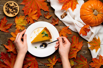 a piece of pumpkin pie on a wooden table with female hands, pumpkin and autumn leaves