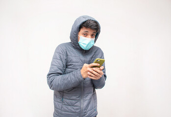 Joven hombre con frio y ropa abrigadora con una mascarilla color azul mientras sostiene un telefono...