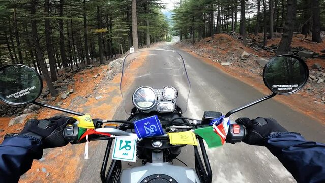 POV Motorcyclist riding motorbike by scenic mountain pine forest in Himalayas. Biker on motorcycle going between mountain valley by landscape path. Steering wheel view with Tibetan buddhist flags