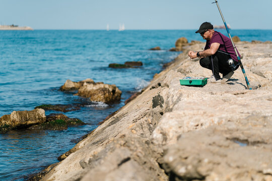 Serious fisherman sitting on haunches on rocky seashore