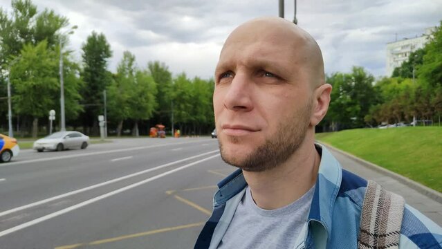 Young man standing near highway, waiting for bus. Cute bald guy at public transport stop. Man in plaid shirt wait for taxi, standing at roadside, looks at road along which cars and trucks traveling