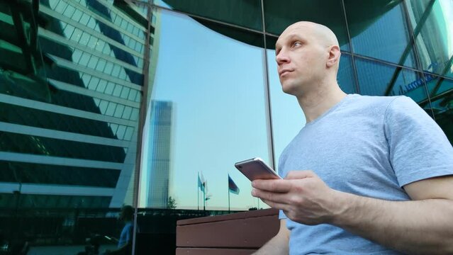 Young man sitting on bench near mirrored skyscraper and using mobile phone. Bald guy with smartphone. Chatting, viewing photos and videos, playing games, surfing the Internet