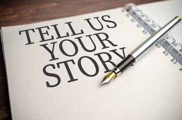 Text TELL US YOUR STORY on paper notepad and pen on wooden background