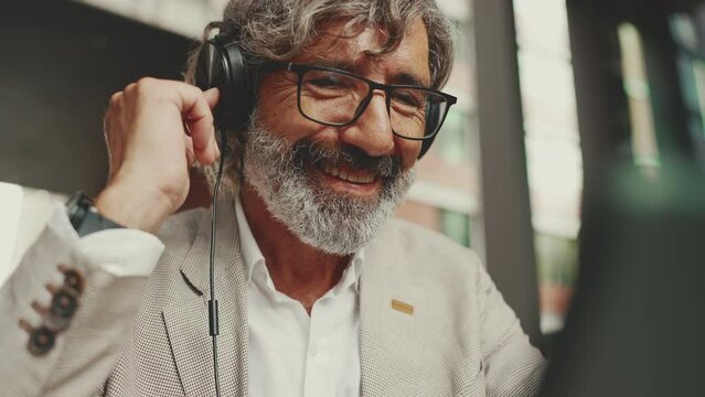 Clouse up, smiling mature businessman with beard in eyeglasses and headphones, sits in an outdoor cafe and works using tablet