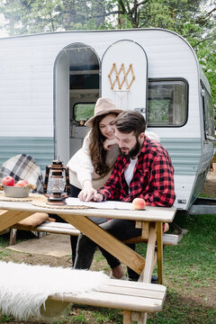 Guy and girl in hat look at tourist maps at the table near the trailer. Happy young caucasian couple traveling in travel van. Romantic atmosphere of relaxation. Road trip around country for weekend