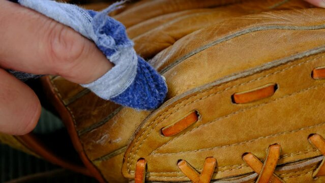 Leather conditioner being applied to an old leather baseball glove by hand with a blue rag, and then being buffed out