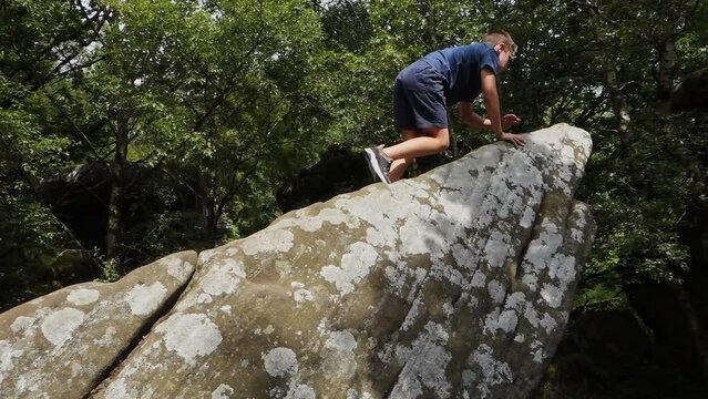 Young boy climbing and jumping on the rocks at Brimham Rocks, once known as Brimham Crags in North Yorkshire, surrounded by woodland in the UK.