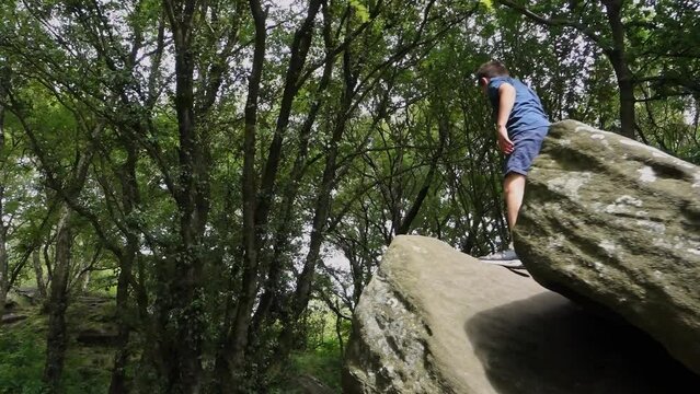 Young boy climbing and jumping on the rocks at Brimham Rocks, once known as Brimham Crags in North Yorkshire, surrounded by woodland in the UK.