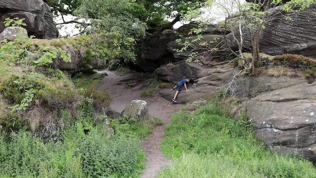 Young boy climbing and jumping on the rocks at Brimham Rocks, once known as Brimham Crags in North Yorkshire, surrounded by woodland in the UK.