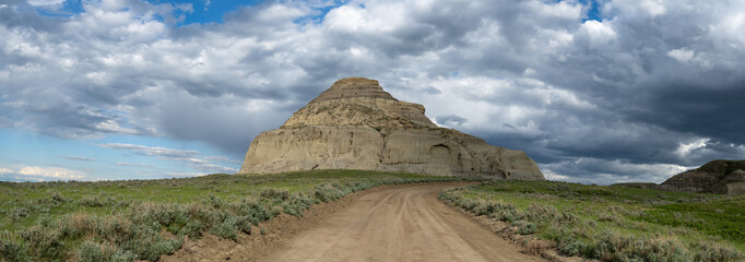 Butte with blue sky with white to gray clouds and a desert foreground including a tan colored dirt road.