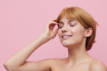 Obraz premium a close horizontal photo of a pleasant, lovely woman with perfect skin, standing on a pink background, gently touching her face with her hands, closing her eyes with pleasure and smiling prettily.