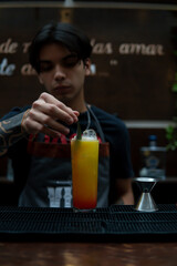Waiter preparing a yellow drink that looks like juice, he is placing a small leaf on it as decoration. The waiter is at the bar counter. Glass of juice with waiter in the background.