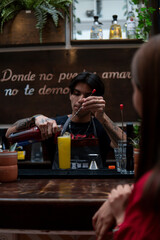 Bartender at the bar counter preparing a drink in front of a client dressed in red with brown hair. Waiter preparing a yellow drink. Girl in a nightclub in front of the bartender.