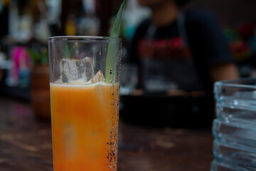 Close up of a glass of cold juice with an ice cube and a green leaf decoration. Drink on a bar counter with the bartender in the background. Yellow drink in a night club.