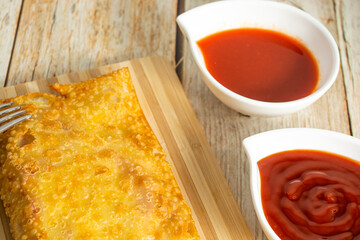 brazilian snack pastel, over wooden table with sauce, pepper, ketchup also fork and knife. traditional pastry