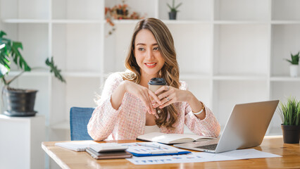Beautiful young smiling Asian businesswoman drinking coffee and using laptop computer at office workplace, feeling happy and relax.