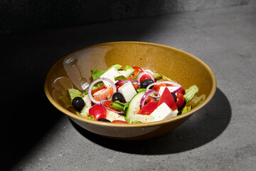 Classic greek salad on ceramic bowl in minimal style. Simple composition with greek salad on gray stone background with hard shadow. Dark concept menu. Vegetable salad on concrete table and shadow.