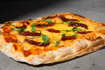 Italian pizza with cheese and dried tomatoes on dark background. Cheese pinsa on gray stone table with hard shadow. Roman pizza with cheese and tomatoes on concrete background. Pinsa menu.
