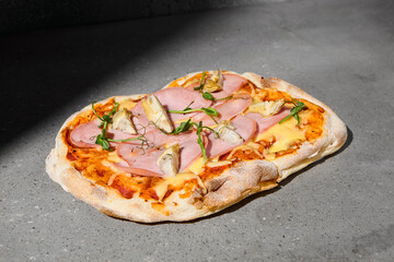 Italian pizza with ham and artichokes on dark background. Ham pinsa on gray stone table with hard shadow. Roman pizza with ham and artichokes on concrete background. Pinsa menu.