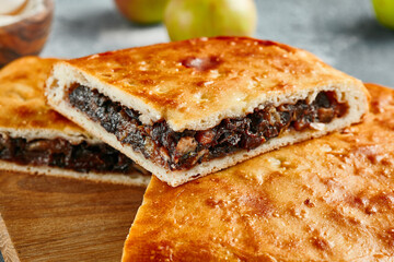 Plum pie on wooden board in rustic style. Aesthetic composition with closed pie with dried plums on concrete background. Sweet pastry - fruit pie with plum and nuts.