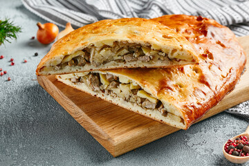 Classic meat and potato pie on wooden board with ingredients. Composition with pork pie on concrete background with textile and spices. Homemade pie with meat and potato in rustic style on gray table.