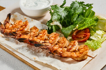 Grilled shrimps with sauce on concrete background. Aesthetic composition with prawn skewer and fresh herbs. Shrimps on grill in summer menu. Seafood skewer in minimal style.