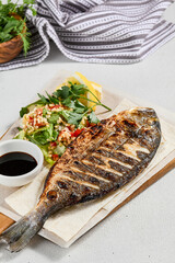 Grilled dorado with garnish on concrete background. Aesthetic composition with bbq fish and vegetables. Healthy food - roasted dorado. Fish dish in minimal style. Whole grilled fish.