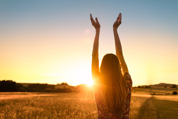Woman standing in an open field with her hands up to the beautiful sunrise feeling energized, and happy. Positive energy, prayer and gratitude concept.