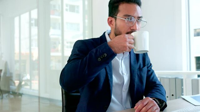 Serious university professor analyzing documents inside a campus office using a computer. Focused male academic researcher enjoying his coffee while typing a thesis or research paper with glasses
