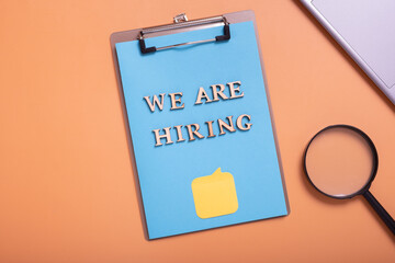 Wooden letters with phrase we are hiring on clip-board with magnifying glass and laptop on colored work table top view. Search worker concept