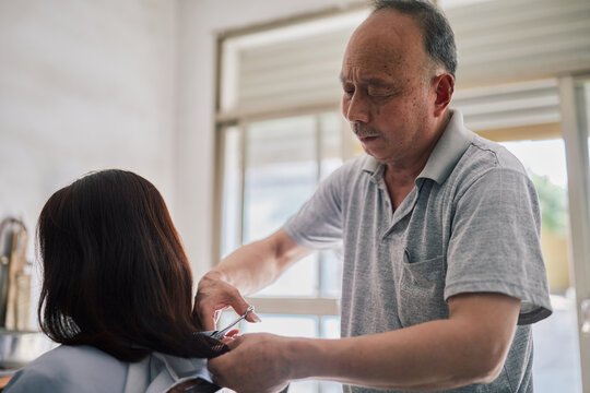 Older Asian Hairstylist Cuts A Customer's Hair At His Salon.